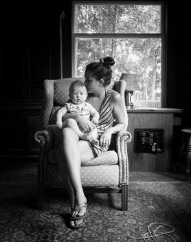 black and white image of thirty something woman holding a baby while sitting in vintage chair by large window.