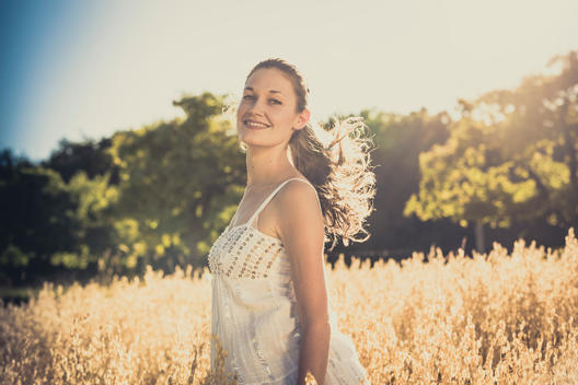 young woman running at summer through a cornfield wearing a light dress and smiling in camera, back light and lens flare