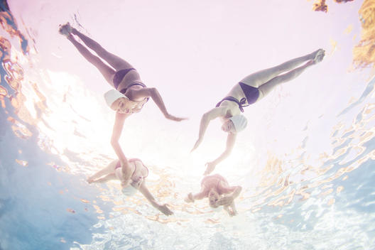 Four synchronized swimmers underwater in white caps and blue bathing suits doing back flips towards each other