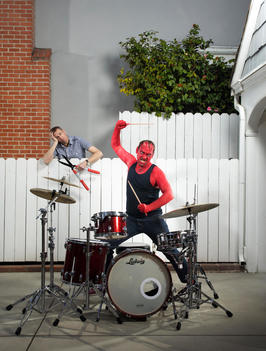A man dressed as a devil plays the drums in his driveway in front of his garage as his neighbor leans on the fence while holding hedge clippers