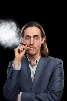 A man with long hair in a suit smokes an e-cigarette