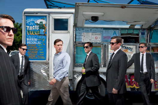At a taco truck a man is surrounded by multiples of himself dressed in suits, exploring our paranoia