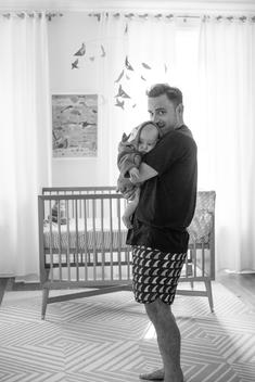 Black & white image of father standing in front of crib holding baby smiling, looking at camera.