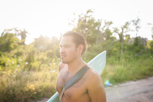 Portrait of muscular, athletic surfer Matt Hernandez with large wings tattoo on chest walking with surfboard on road at Far Rockaway Beach, Queens
