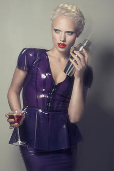 Blonde model, pale skin in a purple dress, red lips, yellow nails, holding a glass shaking a cocktail, looking away