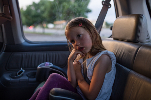 Jane, a young girl with shirt sleeves rolled up, sits cross-legged, impatient and exhausted, in her car seat in the back of her family\'s minivan as they return home. Denver, Colorado