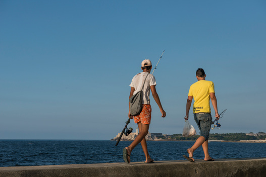 Two young men walking along the top of the Malec?n (Avenida de Maceo), carrying fishing rods. Havana, La Habana, Cuba.