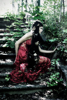 A young woman with long black hair wearing a red night dress crouched on outdoor steps in summer dappled light with leaves on her waiting alone in the undergrowth..