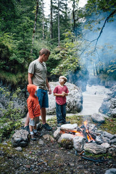 20 something man with 3 year old and 6 year old having campfire by creek in mountainous area in Austria.