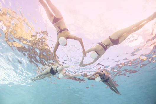 Four synchronized swimmers underwater in white caps and blue bathing suits doing back flips towards each other