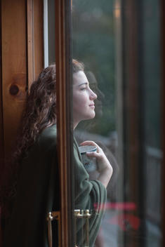 a woman in profile wearing a blanket around her shoulders, holding a mug, leaning against a sliding door looking outside