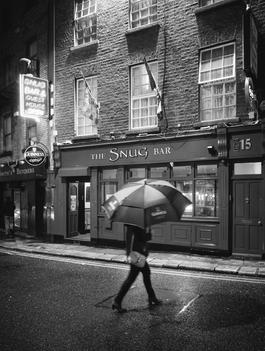 black and white image of woman with umbrella walking on the streets of dublin, ireland at night with bar in the background.