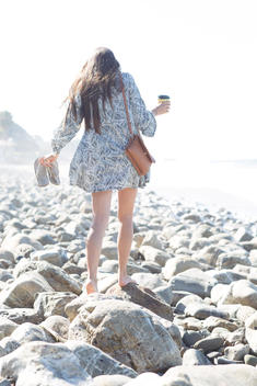 Backlit image of 18 year old brunette girl with long hair in summer dress walking over rocks on rocky beach with purse, flip flops in hand and coffee cup.