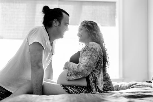 Black and white image of pregnant mom holding belly with father both in pajamas in bedroom with natural light
