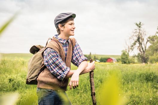 Side view of teen boy in field wearing flat cap leaning against branch looking away smiling