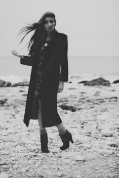 A beautiful women with long dark hair, and a long black coat on the beach.
