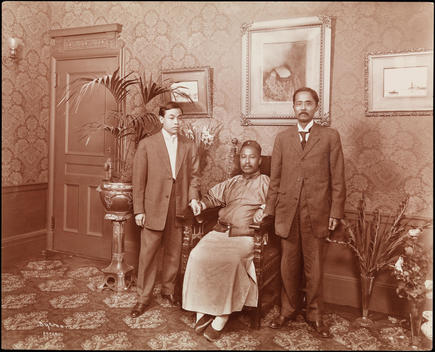Y.S. Wan(S) And Two Male Relatives. The Two Men Are Standing Around The Seated Y.S. Wan(S). They Are Surrounded By Plants And Flowers. He Is Dressed In Traditional Asian Garments And The Other Two Wear Suits. Framed Paintings Hang On The Wall Behind Them.