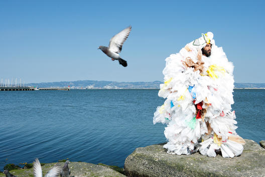 A man covered in plastic bags stands on a rock in front of the ocean as seagulls fly around