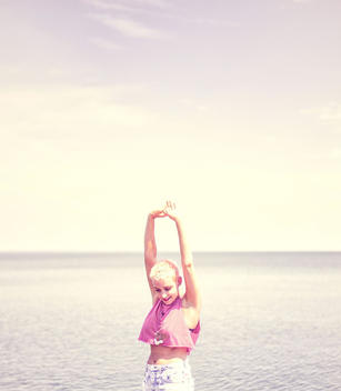 , lake in background. Pink-tinted image of blonde, short haired model stretching her arms