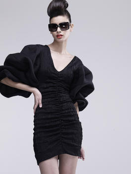 brunette with sculpted hair wearing Oliver peoples sunglasses and Katie Gallagher black couture dress on grey background