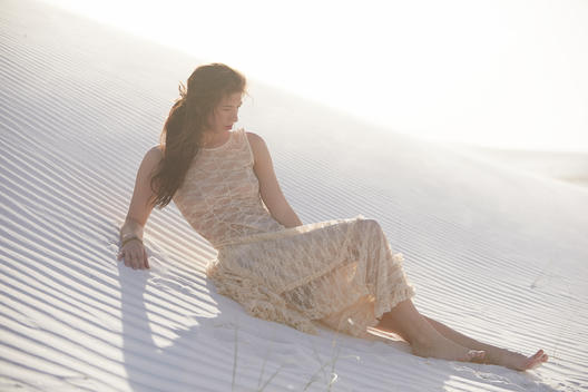 Brunette girl in a long sheer lace dress laying barefoot on the sand dunes while the wind blows