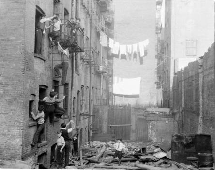 View Of A Space Behind An Apartment Building Filled With Rubble, Children Playing On Fire Escapes, Clothes Hanging On Clothes Lines.