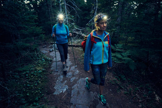 An older woman and a younger woman hiking with headlamps in a dark forest during a backpacking trip.