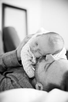 Black & white image of father kissing baby\'s cheek, holding her on his chest on bed,
