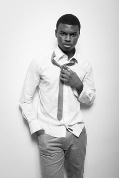 Young black African man wearing a wrinkly button-up white shirt, jeans, and loose tie leans against the wall. New York, NY