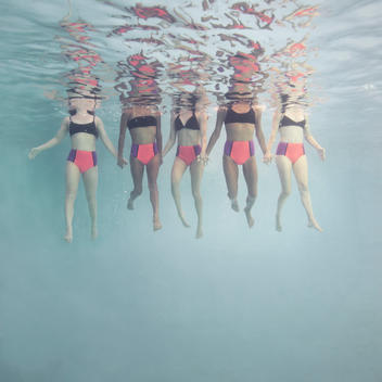 Five synchronized swimmers underwater in a line facing camera treading water with heads out of water