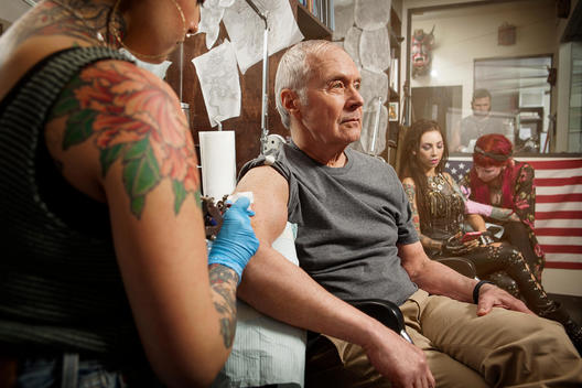 Grey haired senior male getting his first tattoo, by a young female tattoo artist. Another female tattoo artist works on a young female customer in the background.