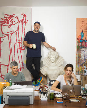 Art dealer, Stefan Simchowitz stands on his desk in his office surrounded by works of art and his assistants working at a desk in front of him