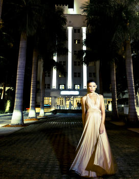 A sexy girl in evening wear in front of a hotel with neon lights and palm trees.