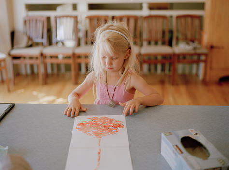 A young blonde girl looks at a paper with the bloody outline of a human placenta on the kitchen counter which her mother is preparing by drying and encapsulating the placenta. The resulting placenta capsules will be ingested by the woman to whom it belong