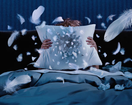 Woman In Bed Sneezing Into A Pillow Creating A Hole And Flying Feathers.
