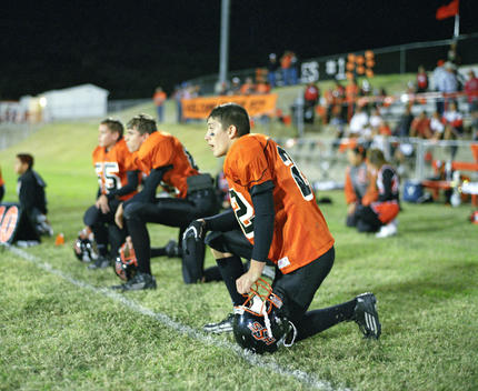 Kneeling High School Football Players On Sideline At Nighttime Football Game.
