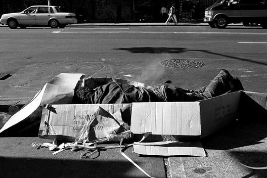 A man sleeps in a large packing box laying on the sidewalk during the day.