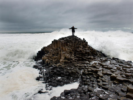 Man standing in a storm atop Giants Causeway getting sprayed by wind, waves and rain.
