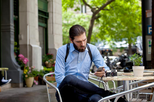 Young man with beard looking at phone sitting at street cafa