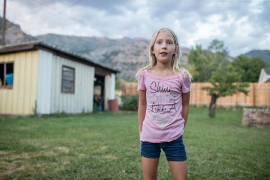 Mabel, a young blonde girl wearing a shirt that says Shine Bright Like a Diamond, stands alone in the backyard, waiting for a new round of a running game she\'s playing with other children, at sunset on a summer evening with the Wasatch mountains and run-d
