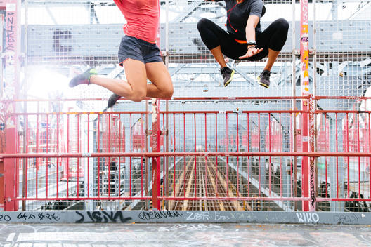 The legs of two women wearing workout clothing, leaping in the air on an urban bridge.