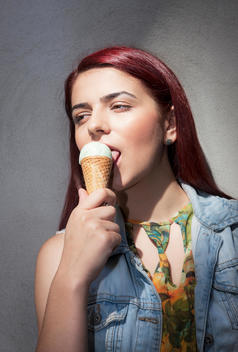 A young red headed woman leans confidently against a wall while licking and icecream cone on a summers day