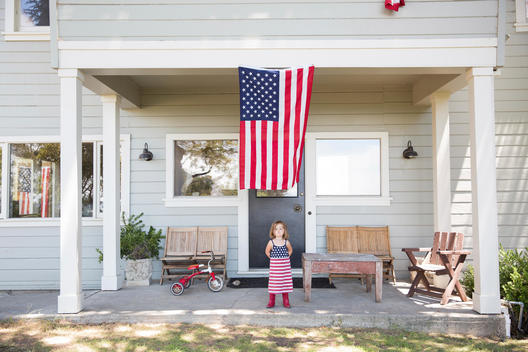 toddler girl standing in front of house with american flag hanging and wearing american flag dress and red boots