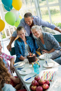 A birthday party in a farmhouse kitchen. A group of adults and children gathered around a chocolate cake.