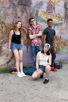 Four teenage friends, 1 black male teen 1 white male teen and 2 white female teens, in front of a graffiti wall in Coney Island Brooklyn, summer group shot