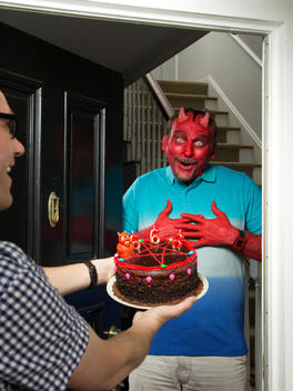 A man dressed as a devil holds his hands to his chest as he excitedly looks at a cake that his neighbor is handing him at the front door