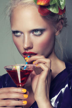 Portrait of young blonde model, wearing bright make up, fruit in her hair, sipping on a cocktail