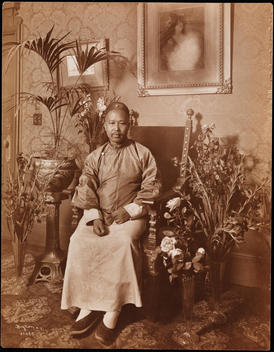 Mr. Wan(S) Sitting In A Chair, Surrounded By Plants And Flowers. He Is Dressed In Traditional Asian Garments. Framed Paintings Hang On The Wall Behind Him. The Envelope Notes The Place As The Hotel Astor.