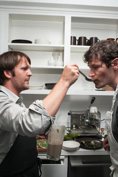 Chef Ren_ Redzepi feeding chef Daniel Patterson with a spoon in a kitchen