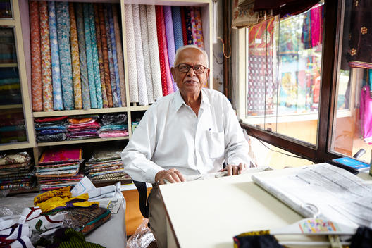 An Indian man sits inside of his fabric shop.